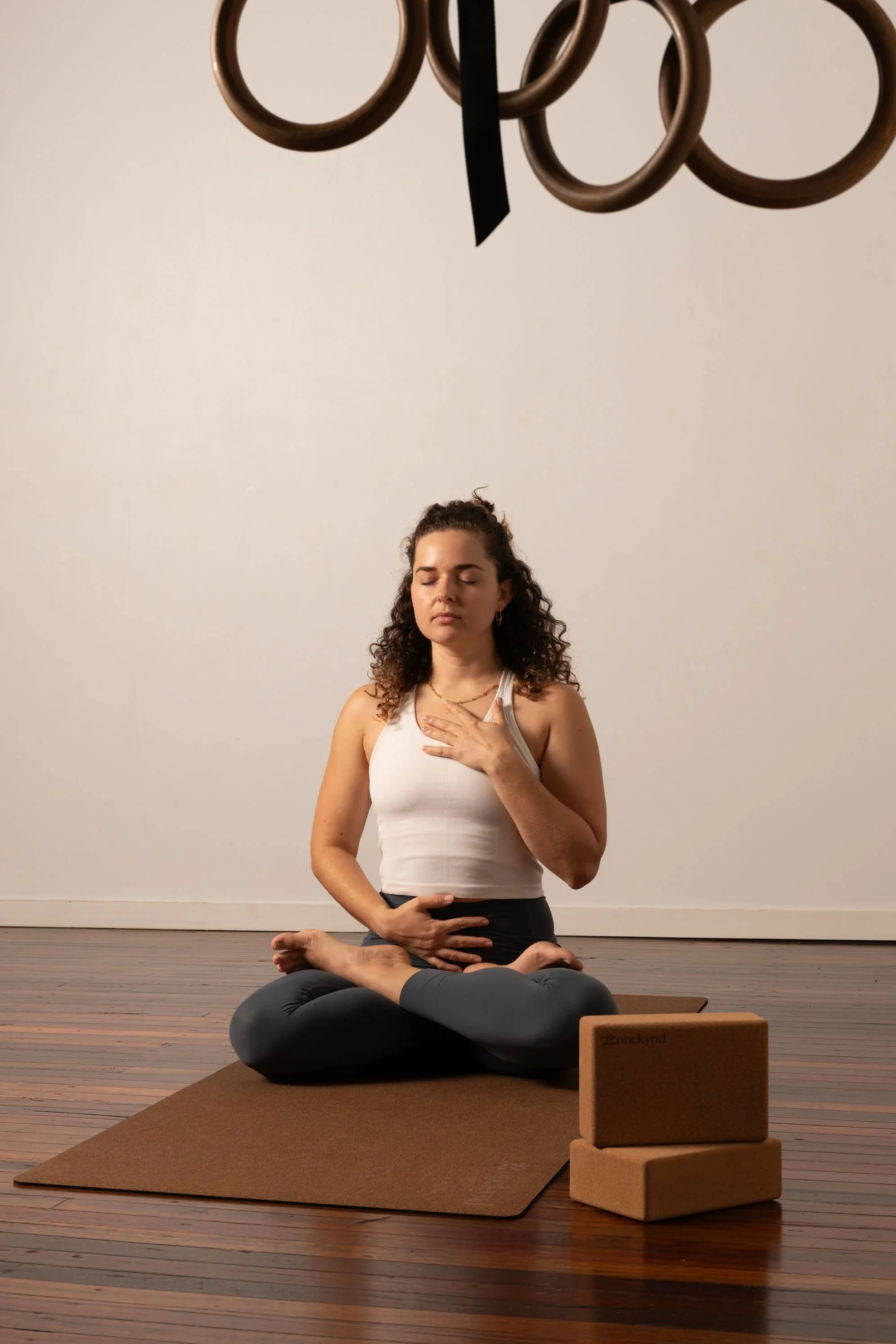 A woman practicing yoga in a seated meditation pose on a yoga mat, with her eyes closed, one hand on her chest, and the other hand on her stomach, inside a room with wooden floors and white walls, alongside yoga blocks.