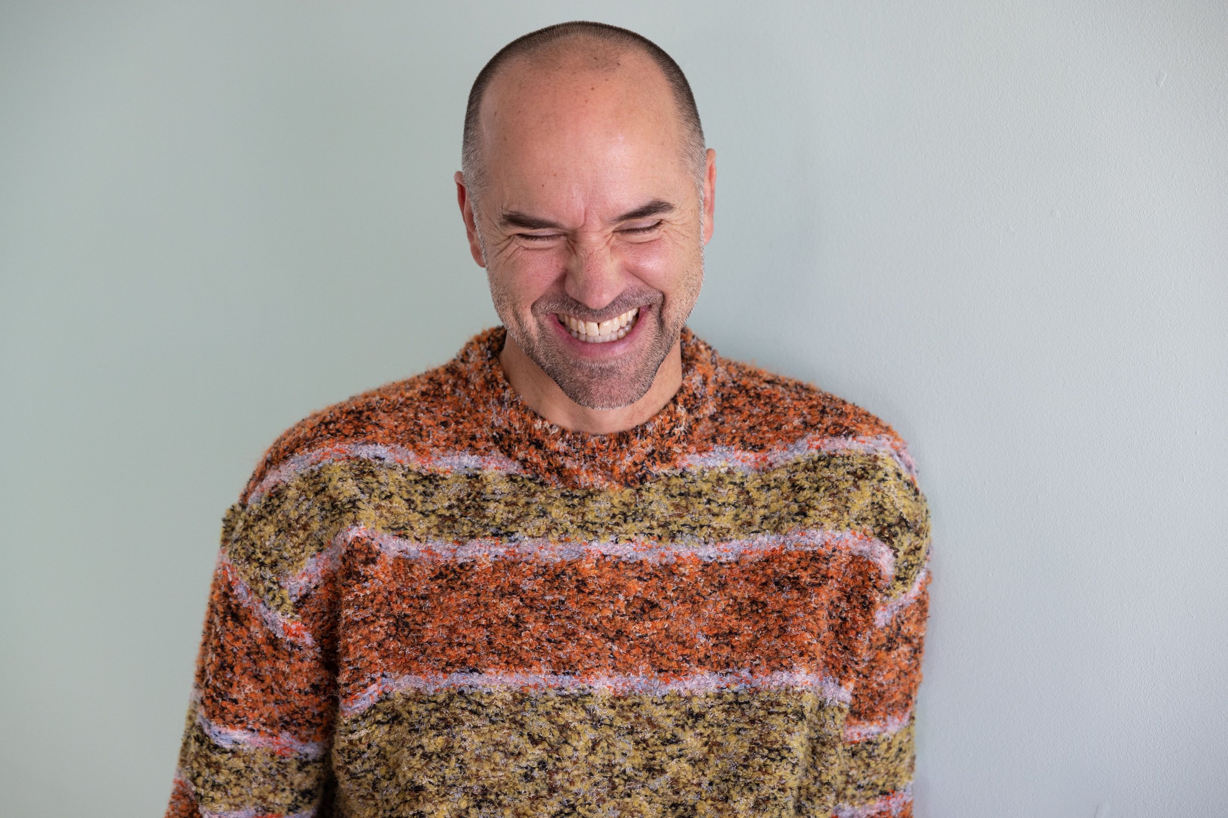 A smiling man with a shaved head and gray beard, wearing a multicolored striped textured sweater, standing against a plain light green wall.