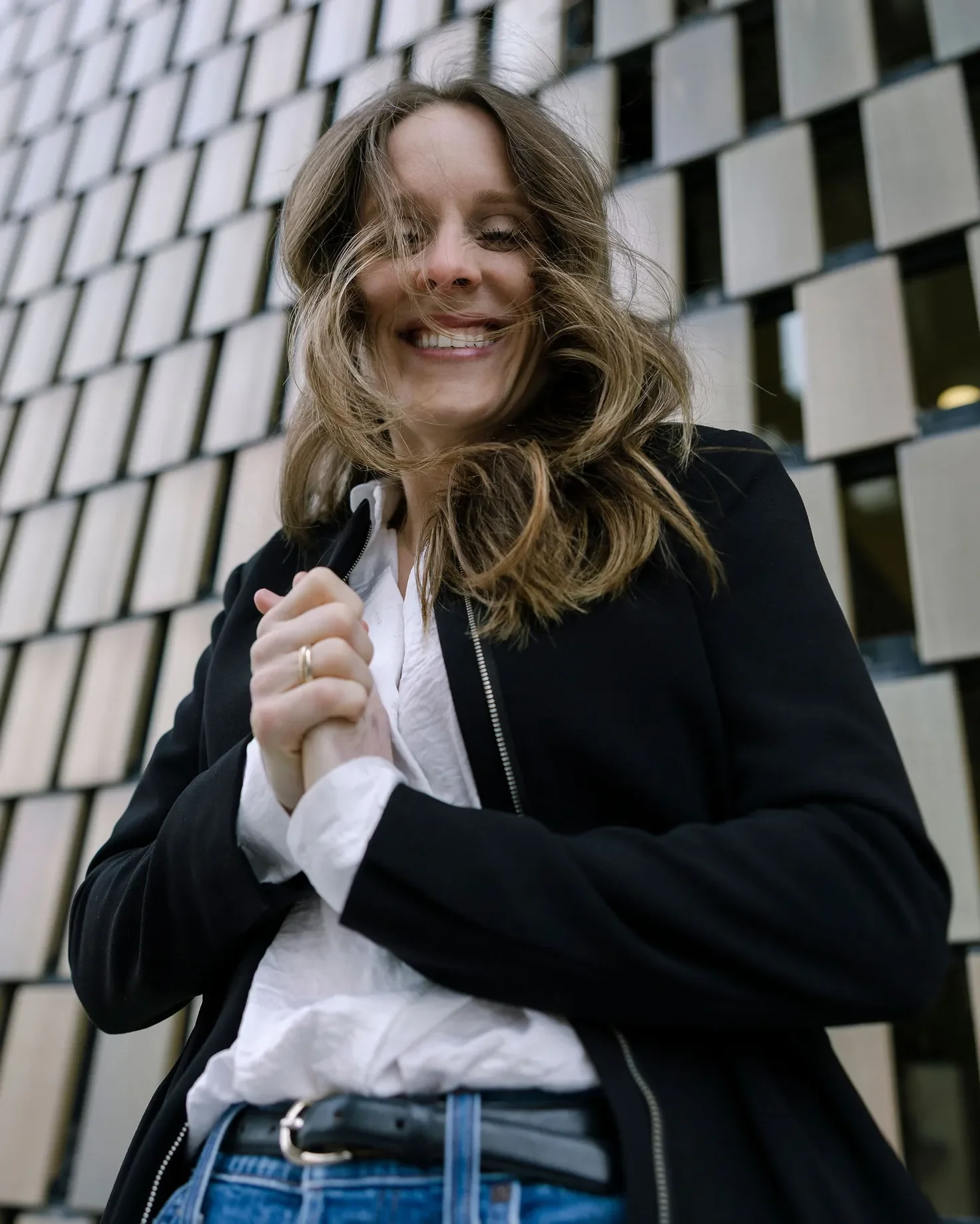 A woman with long wavy hair smiling and looking down, holding her hands together in front of her chest, standing in front of a modern building with square windows.