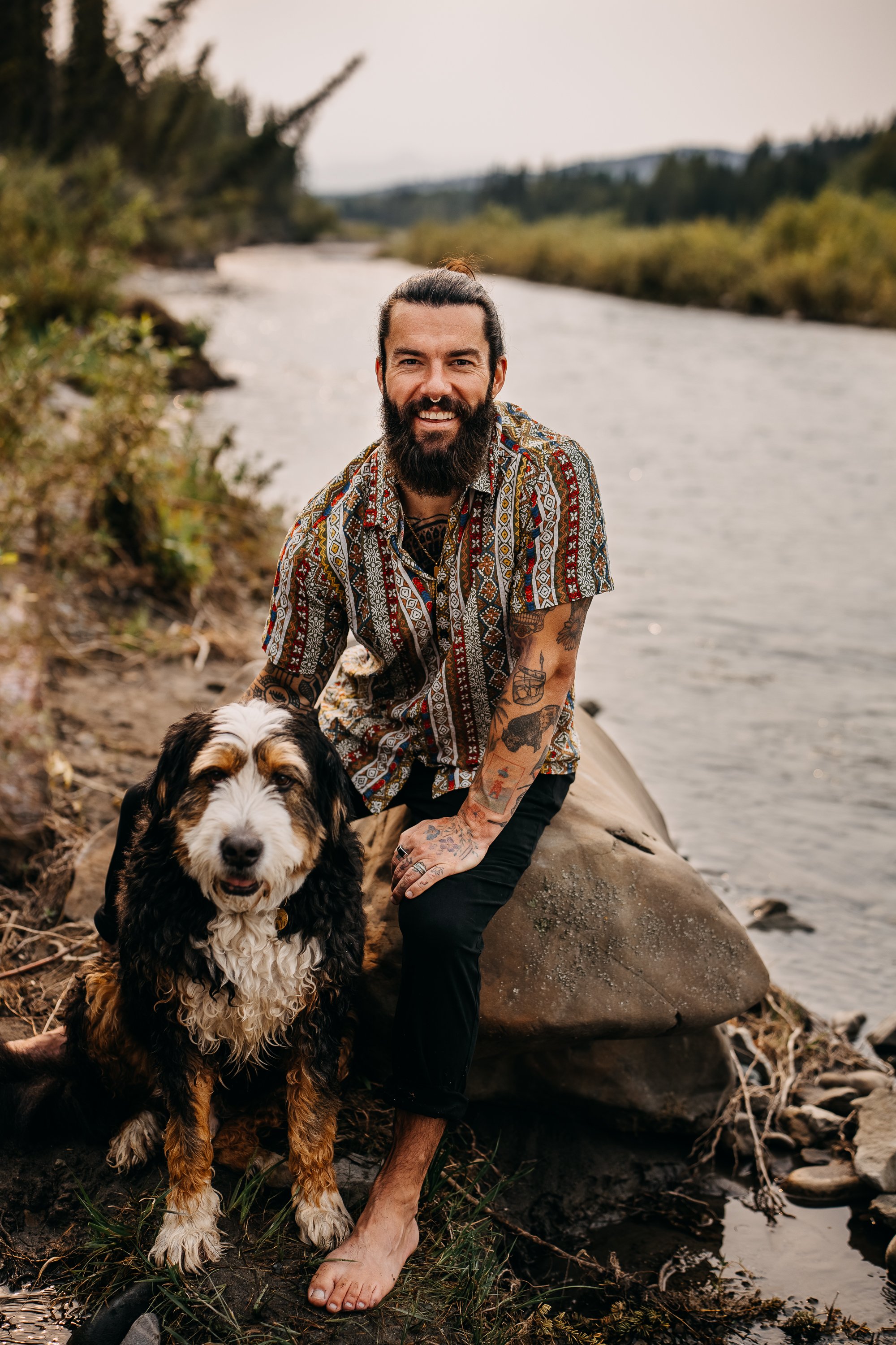 A man with a beard and tattoos wearing a colorful shirt and black pants is smiling while kneeling on a rock by a river, holding a dog on a leash.