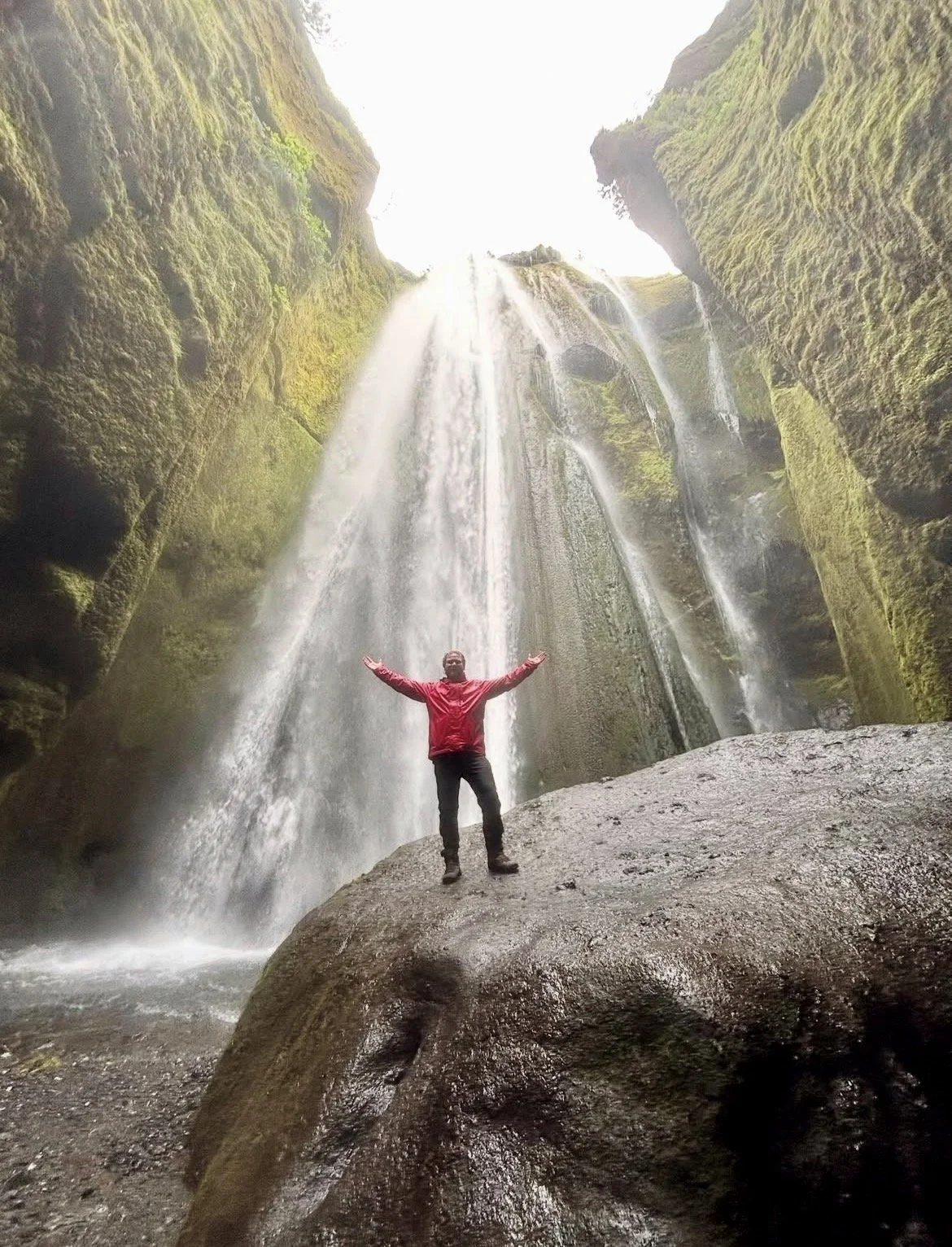 Person standing with arms outstretched on a wet, large rock in front of a tall waterfall surrounded by moss-covered cliffs.