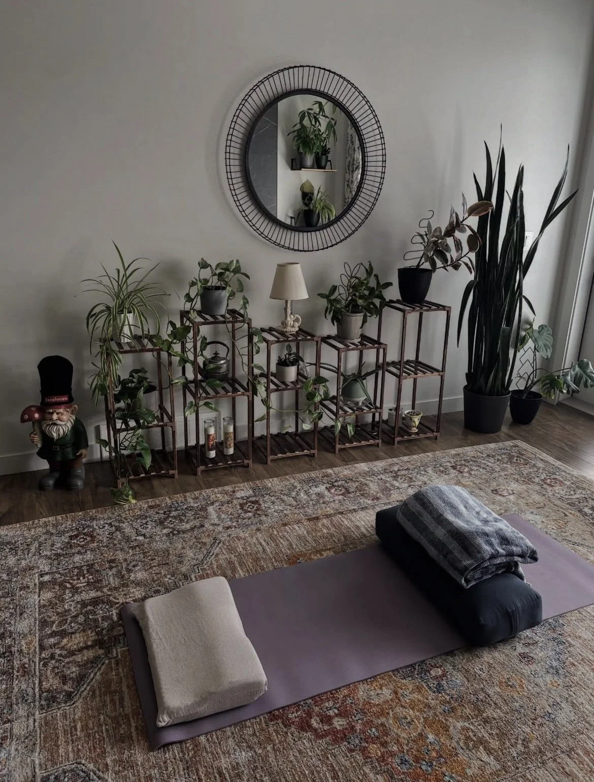 Indoor scene with a yoga mat and rolled towels on an ornate rug, and a shelving unit with potted plants, a lamp, and candles. A large mirror with a decorative frame hangs on the wall above the plants, and a tall houseplant stands nearby.