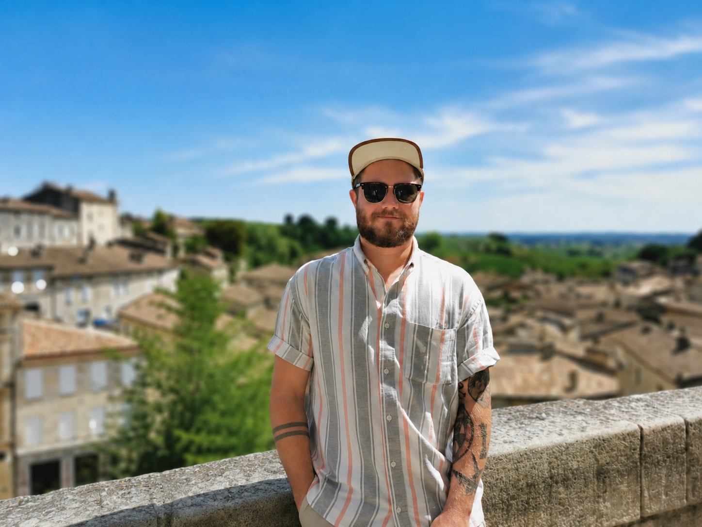A man wearing sunglasses, a baseball cap, a striped short-sleeve shirt, and tattoos on his arms stands outdoors next to a stone wall, with a scenic view of rooftops and green landscape under a blue sky in the background.
