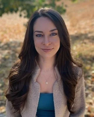 A young woman with long dark hair, wearing a beige jacket and blue top, standing outdoors with a blurred background of trees and sunlight.