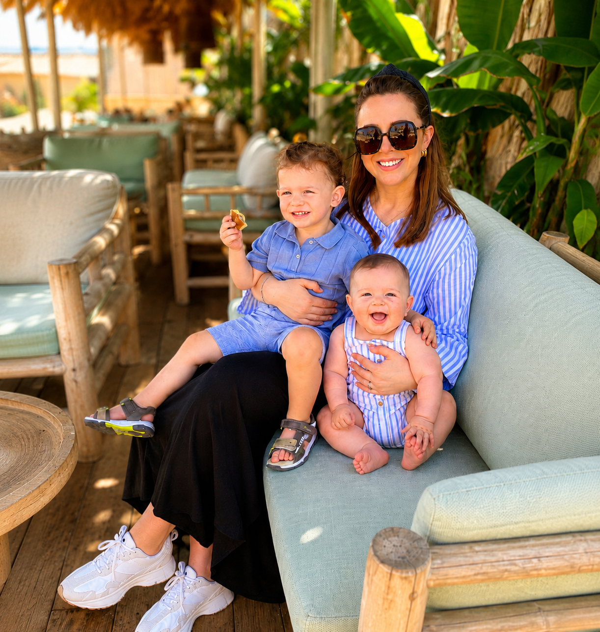 A woman wearing large sunglasses and a blue striped shirt sitting on a light green outdoor sofa, holding two smiling young children in blue and white striped outfits in a sunny patio with green plants and wooden furniture.