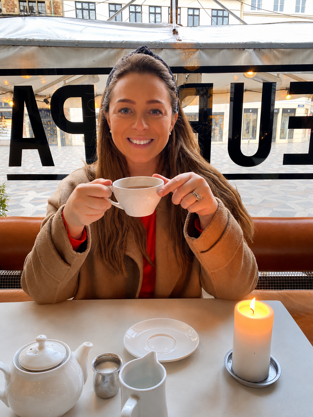 A woman with long brown hair and a beige coat sitting at a table in a restaurant, holding a white cup of coffee or tea, smiling at the camera. There is a white teapot, a small metal pitcher, a white creamer, and a lit candle on the table, with large windows behind her showing an outdoor plaza.