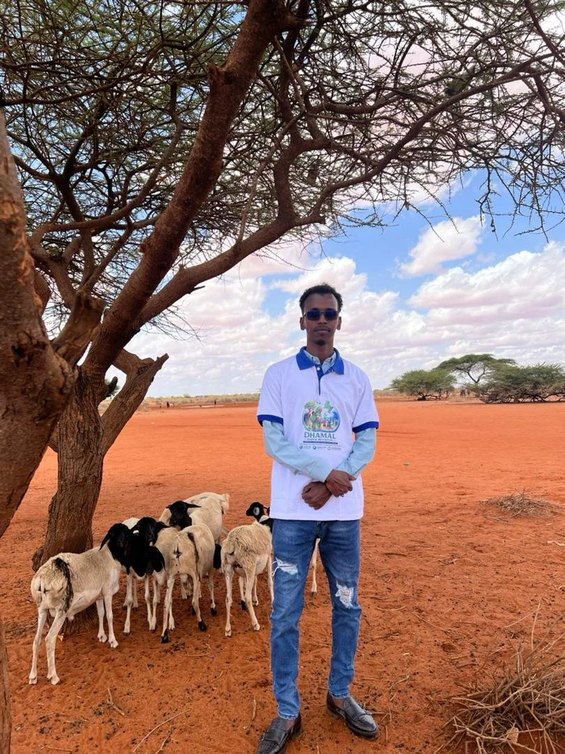A man wearing sunglasses and a white and blue shirt standing on red dirt ground under a tree with a flock of goats nearby, with many trees and a partly cloudy sky in the background.
