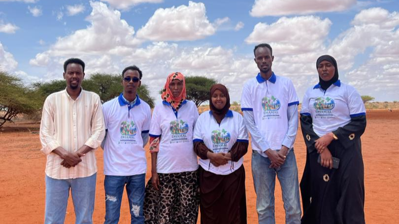 Six people standing outdoors on red dirt ground under partly cloudy sky, some wearing white shirts with a logo and text, others in casual or traditional clothing.