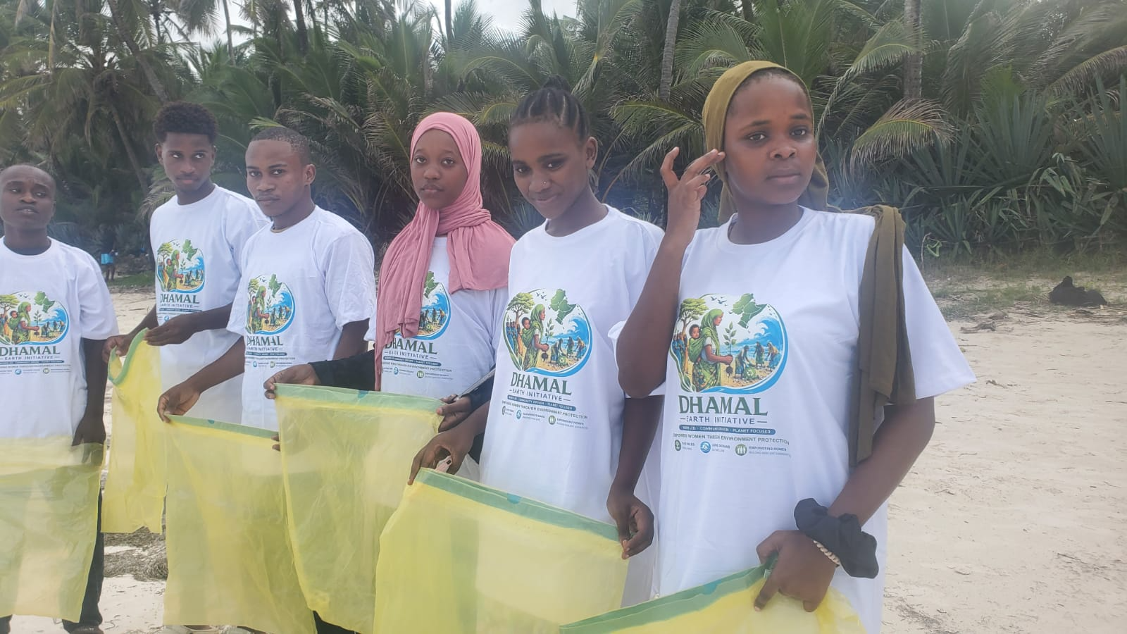 Group of young people standing on a beach holding yellow recycling bags, wearing white t-shirts with a logo for the Dhamal Earth Initiative, with palm trees in the background.