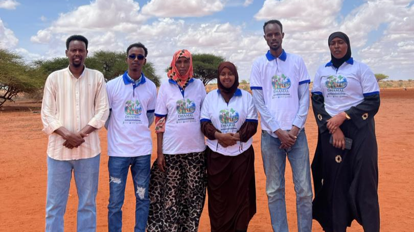 Six people standing outdoors on a reddish-brown dirt field with trees and blue sky in the background, some wearing white T-shirts with a logo and text.