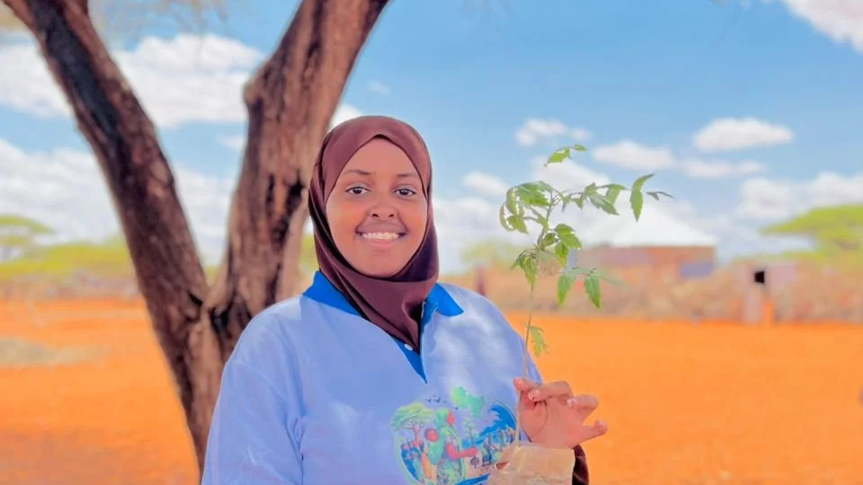 A woman in a blue shirt and brown hijab holding a small plant outdoors in a sunny environment with trees and a brick building in the background.