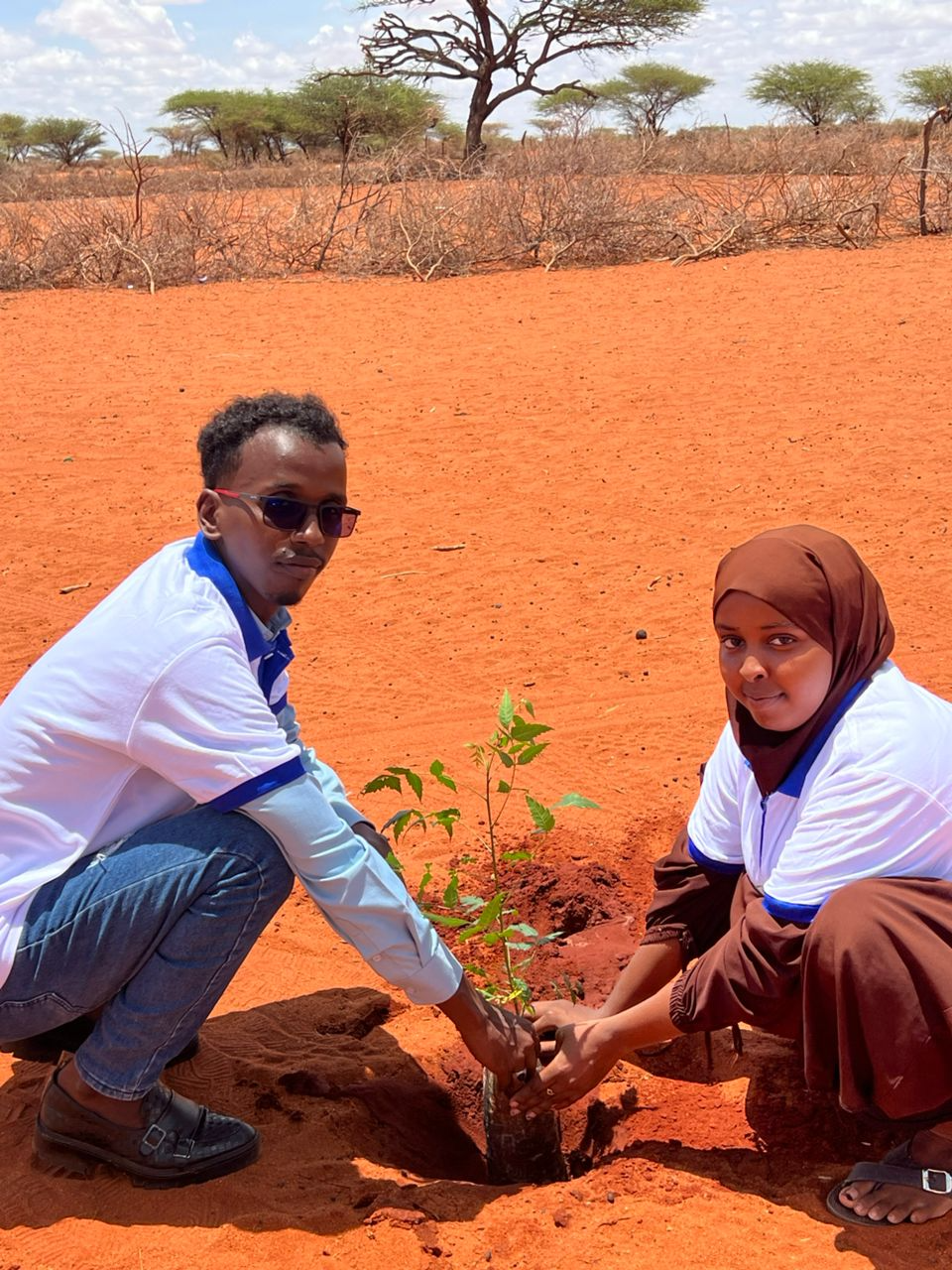 Two young people planting a tree in a dry, sandy landscape with sparse trees in the background.