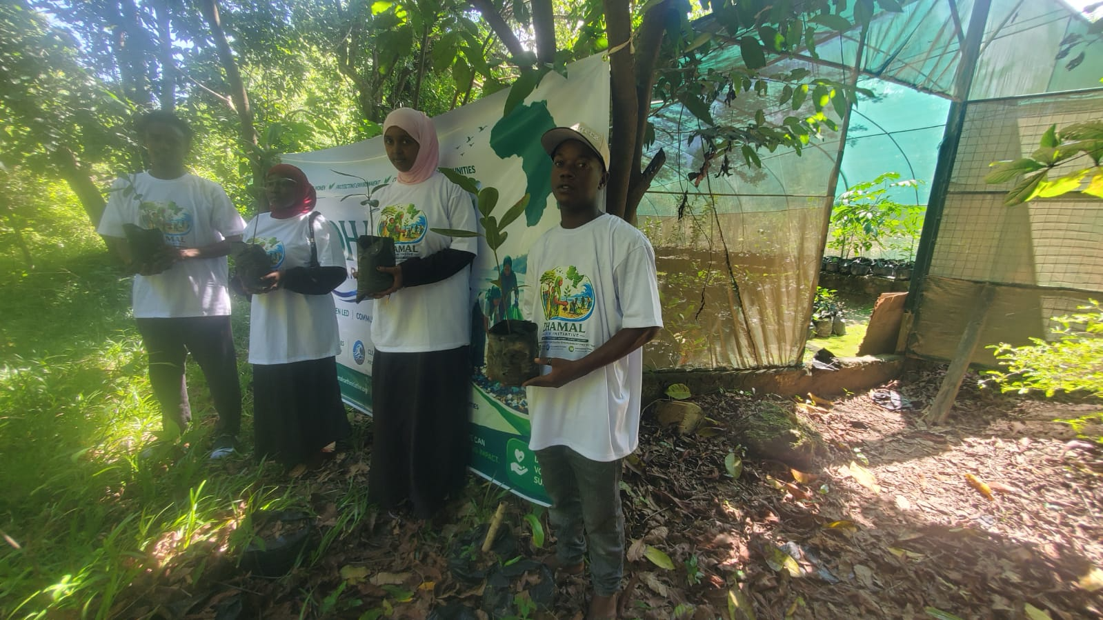 Four young people standing outdoors in a wooded area, each holding a small potted plant, participating in an environmental initiative event with a banner behind them.