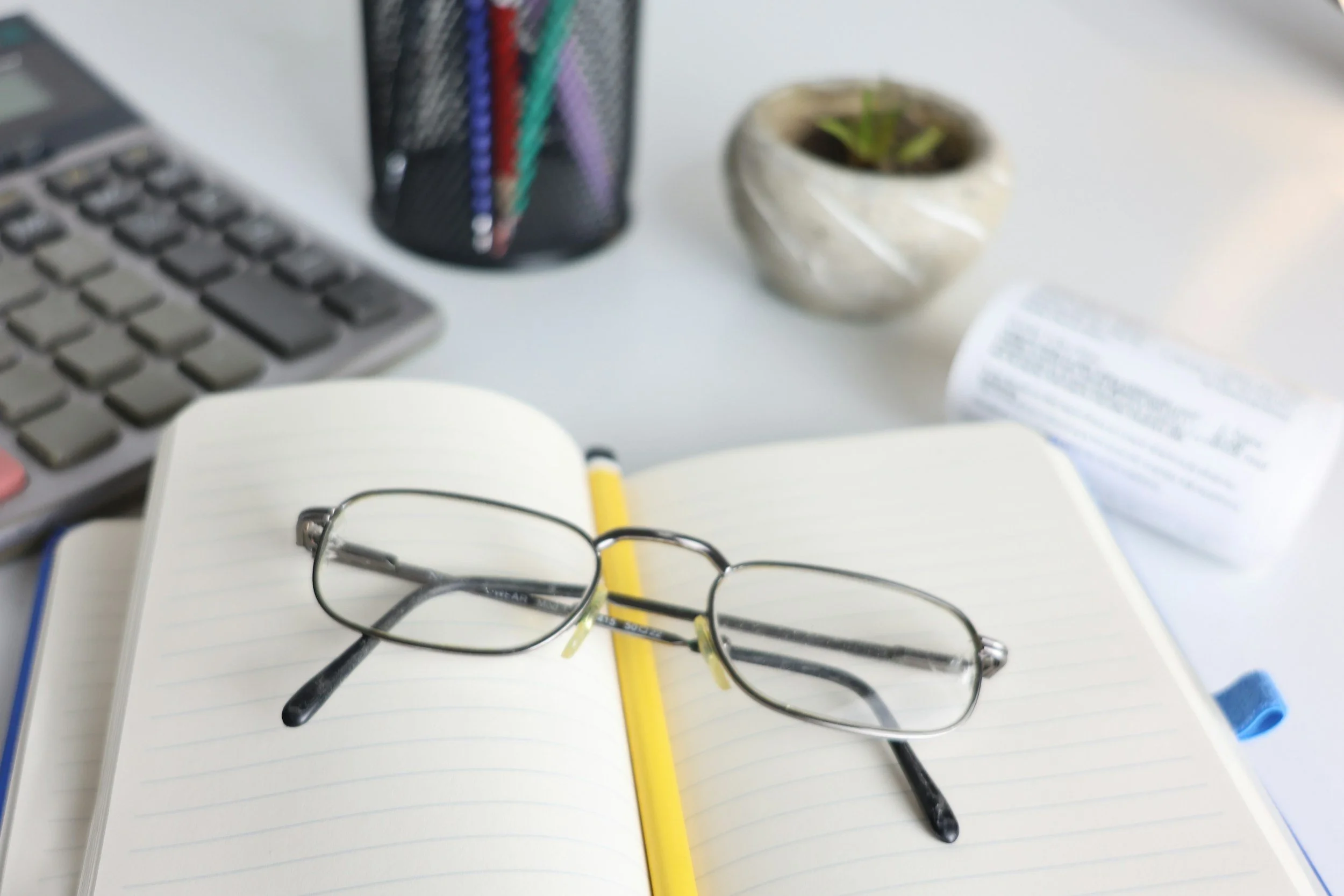 A desk with a pair of eyeglasses on an open notebook in the center view,  and in the background there is a gray keyboard, a black pen holder with colorful pens, a small potted plant, and a partially visible printed document.