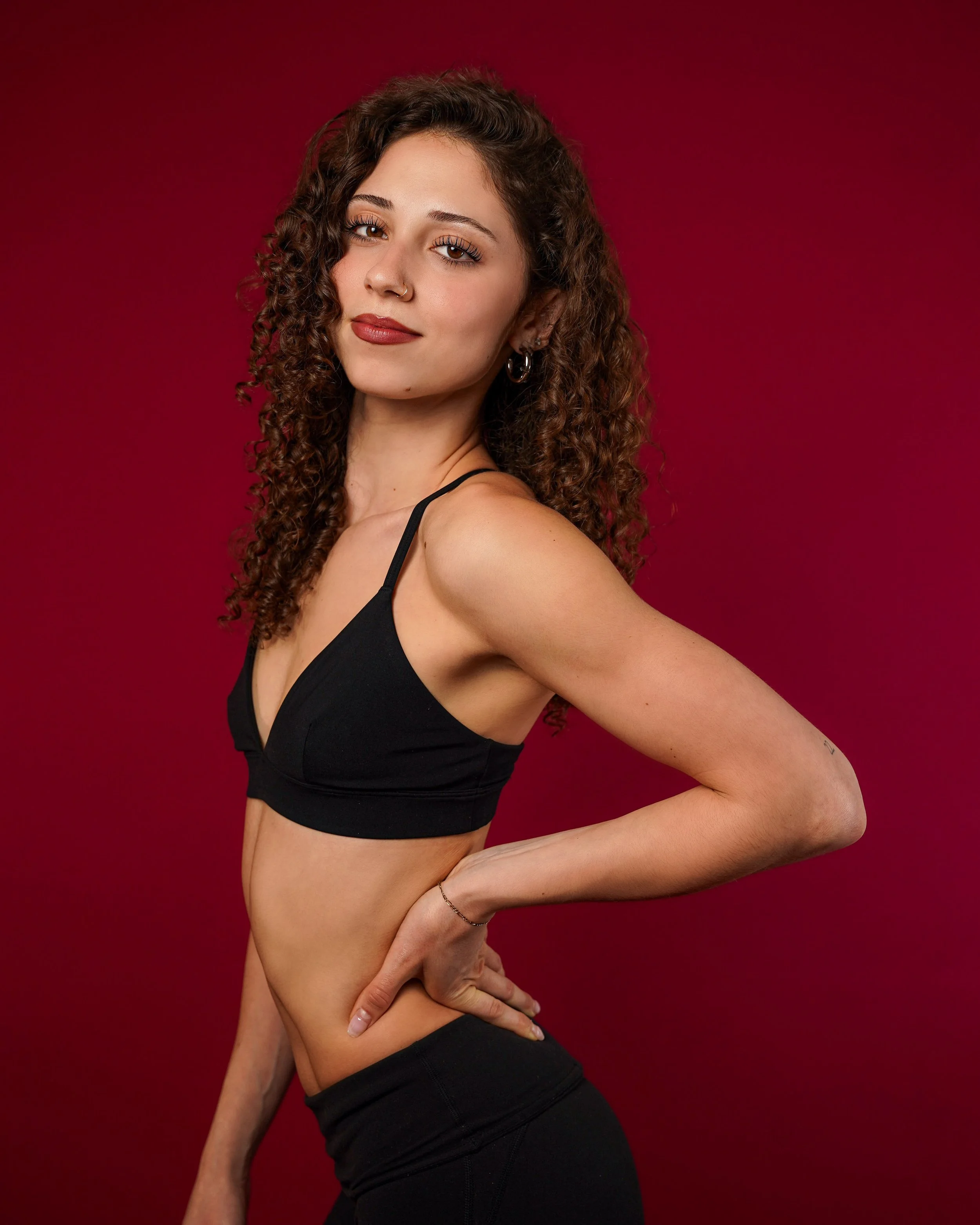 Portrait of a young woman with curly brown hair, wearing a black top, posing against a red background.