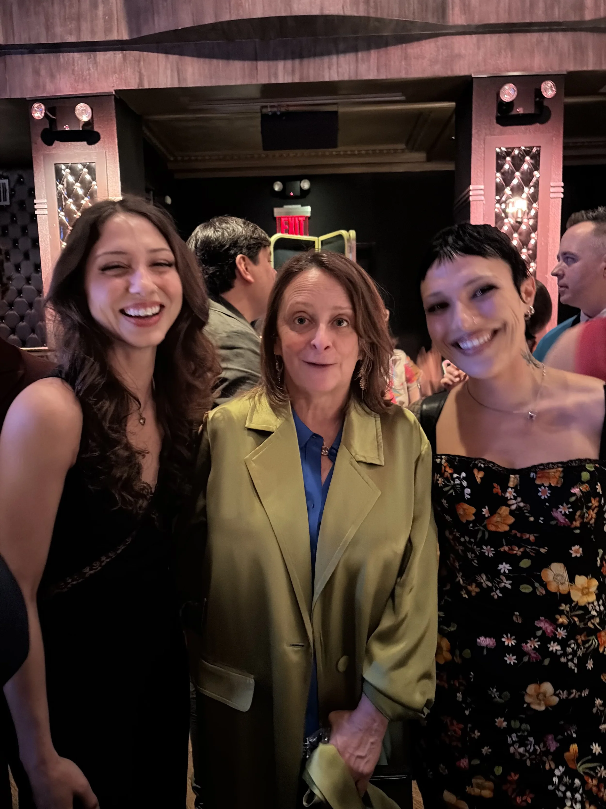 Three smiling women at a social gathering, standing close together indoors with a dark background and patterned walls.