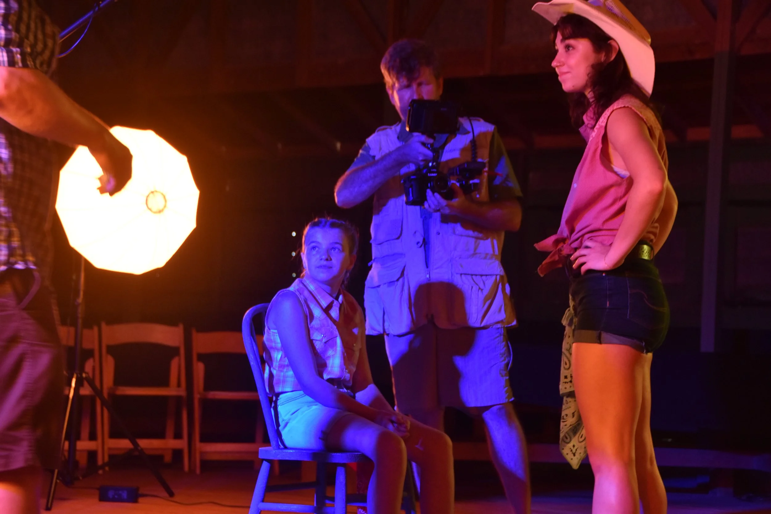 A woman wearing a cowboy hat, a sleeveless top, and shorts is standing with her hands on her hips, looking at a girl sitting on a chair. The girl has braided hair, is wearing a plaid shirt and shorts, and is looking up at the woman. A person holding a camera is filming in the background. The scene appears to take place in a barn or wooden structure with warm lighting.