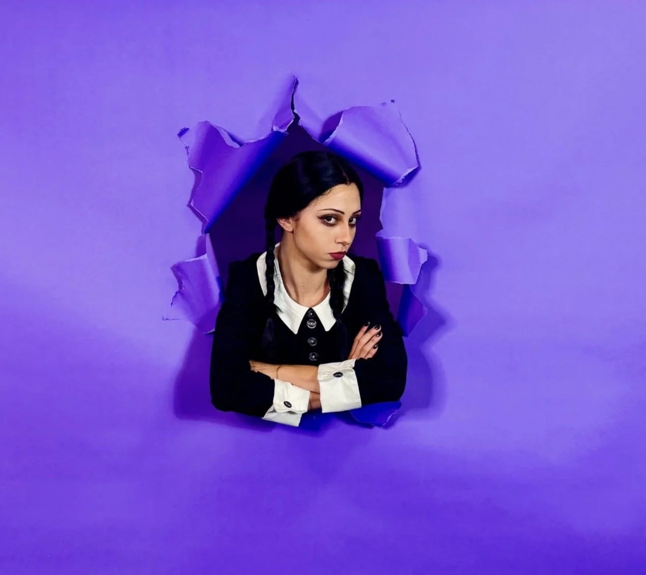 A young woman with dark hair and dark makeup, dressed in a black and white outfit, appears to be emerging through a torn purple paper backdrop, with her arms crossed and a serious expression.