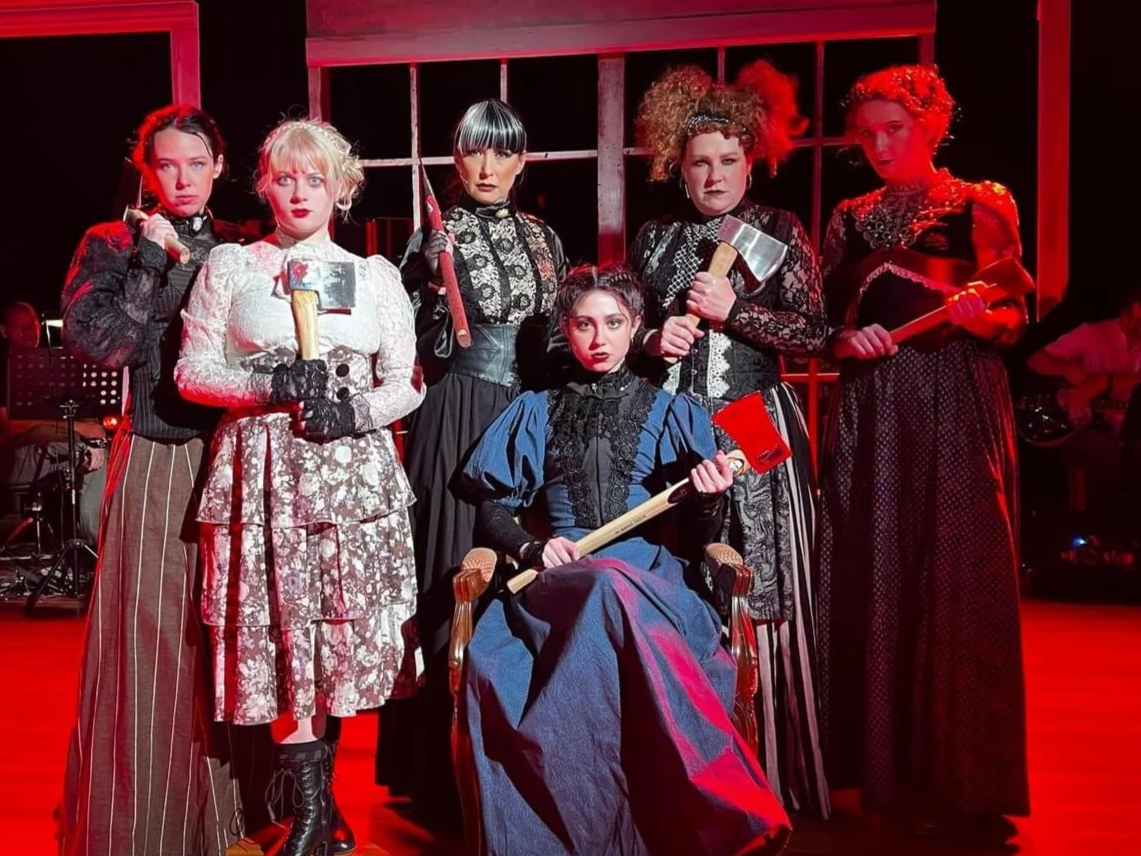 Six women dressed in vintage, Victorian-style costumes, holding various weapons, posing in a theatrical setting with red lighting.