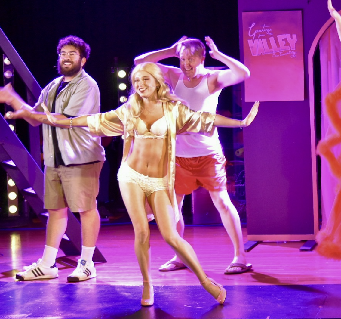 Three performers on stage, one woman in lingerie and satin robe dancing, two men in casual summer clothes, smiling and posing. Stage lights and a sign in the background reading 'Greetings from Valley'.