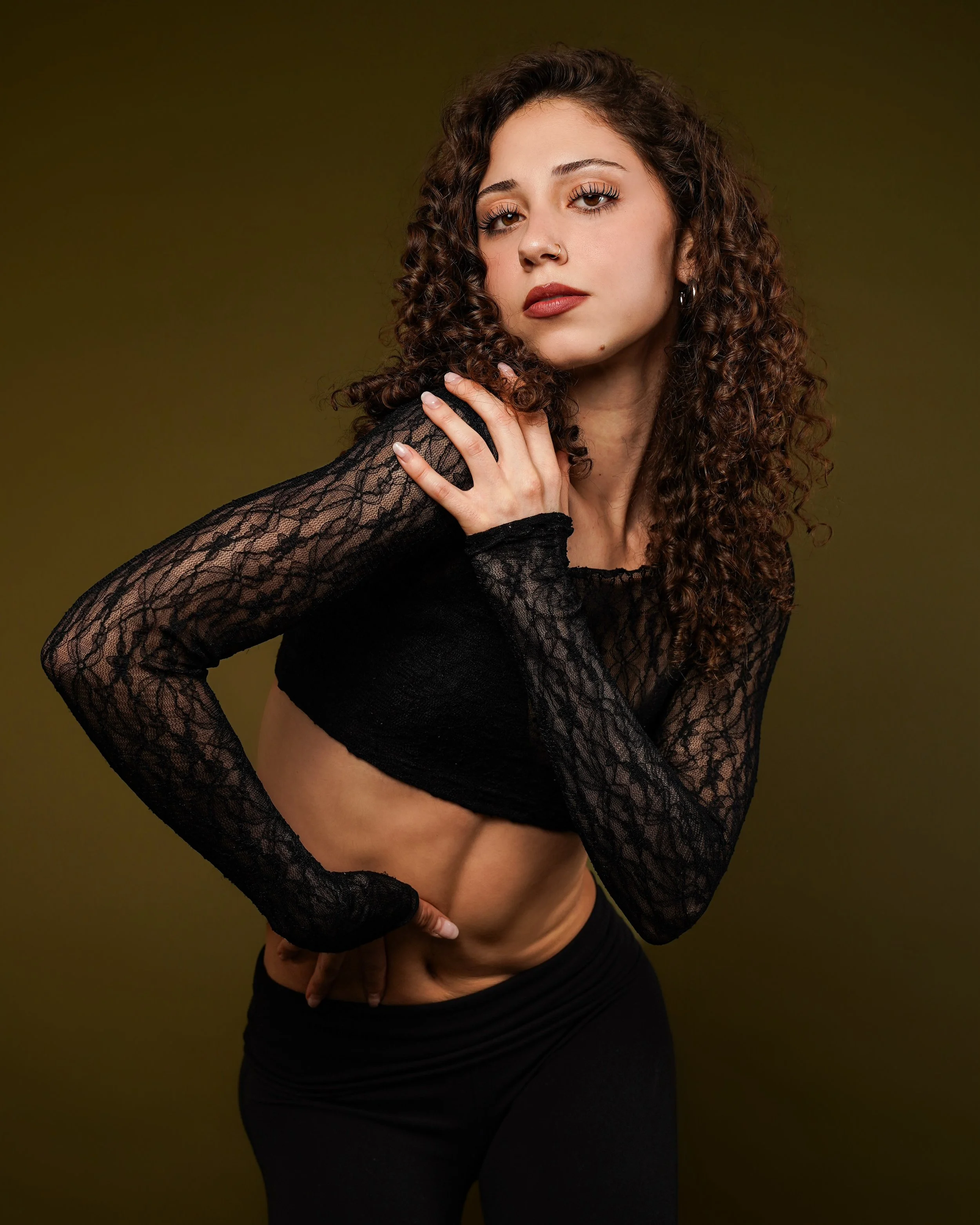 A young woman with curly brown hair wearing a black lace long sleeve top and black pants, posing with one hand on her shoulder and the other on her hip, against a dark green background.