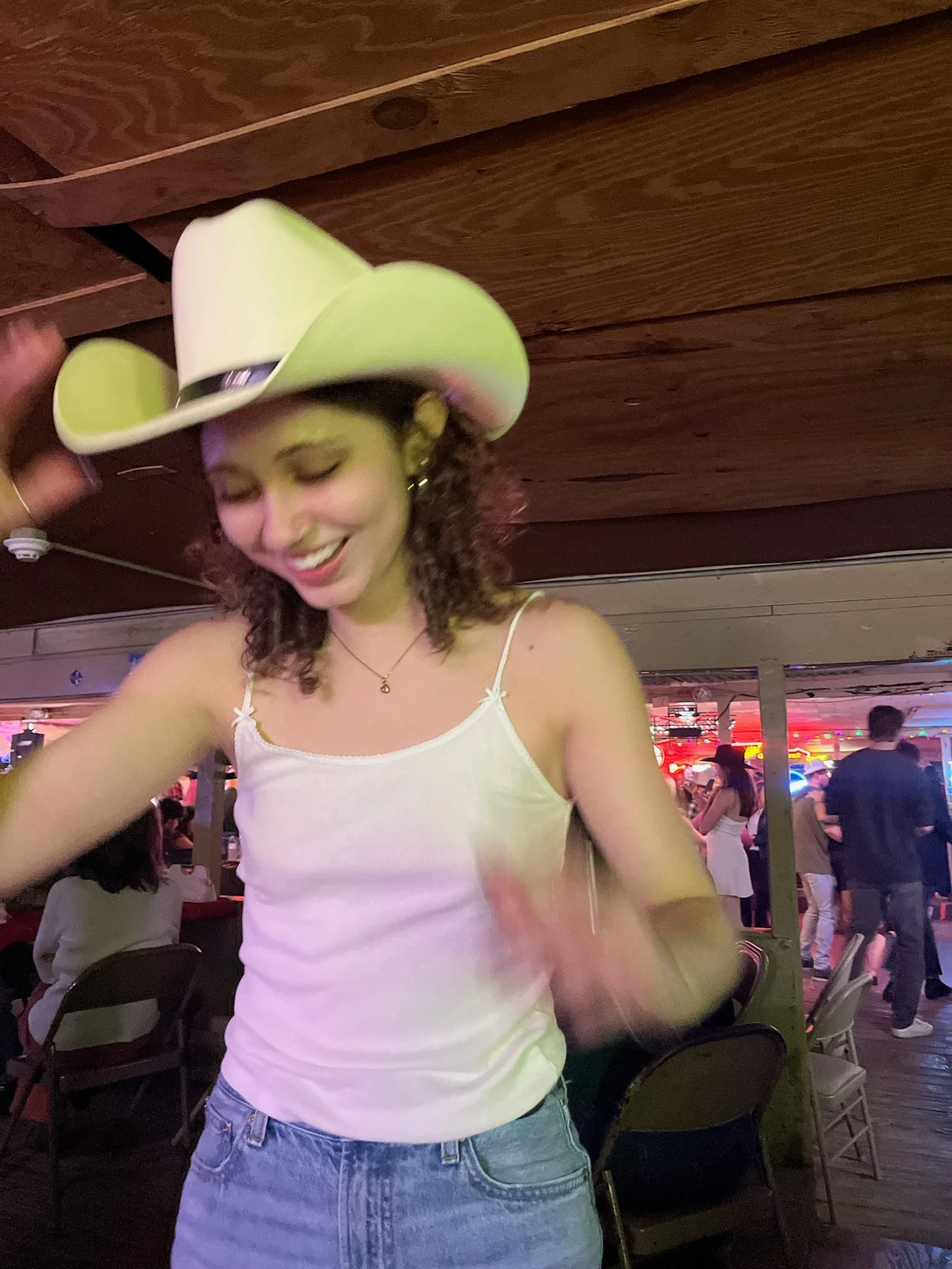 A young woman with curly hair wearing a white tank top, blue jeans, and a large white cowboy hat, smiling with her eyes closed in a lively indoor setting with people and colorful lights.