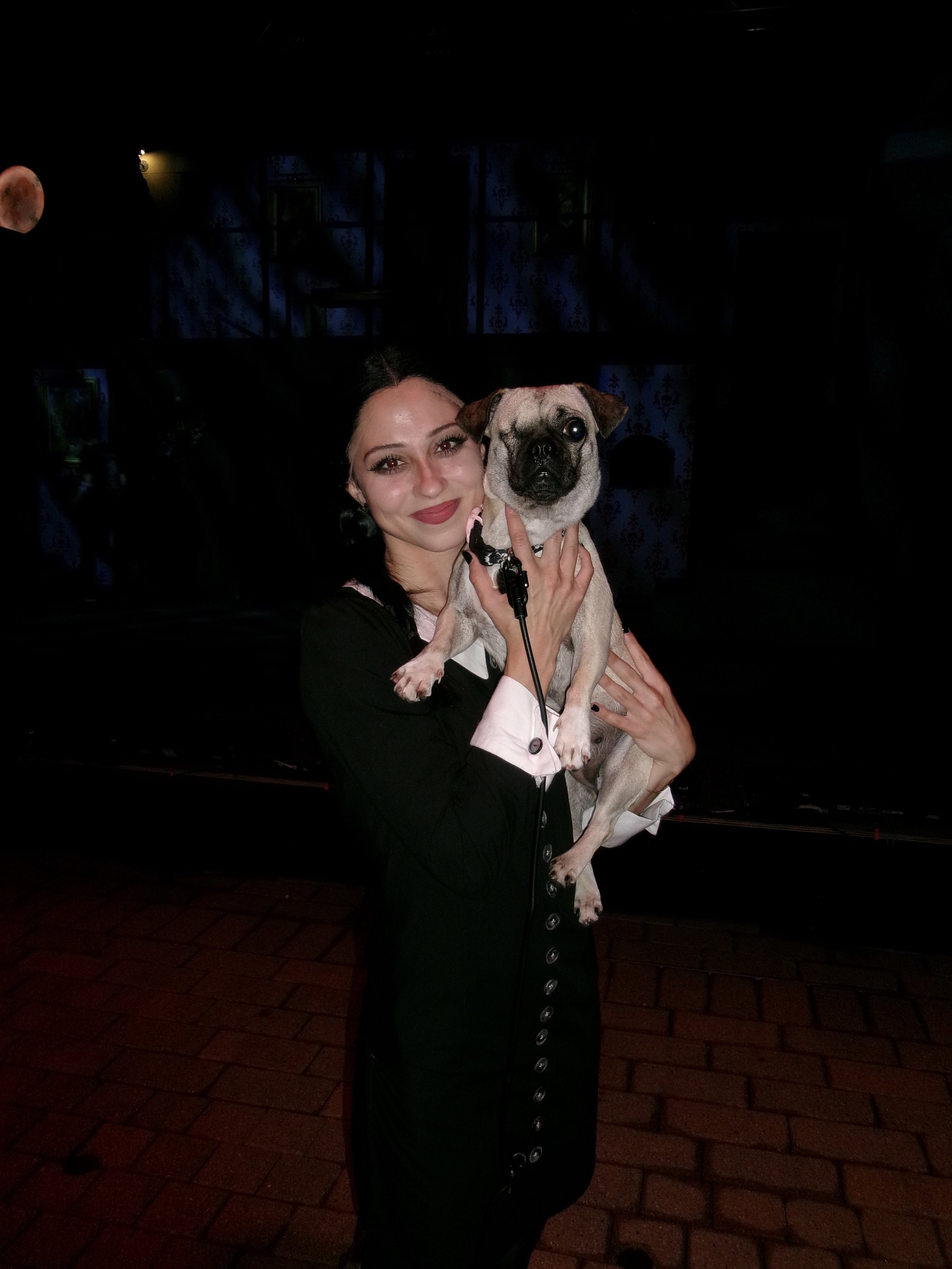 A woman with dark hair and makeup holding a small pug dog with a leash in front of a dark background with moon imagery.