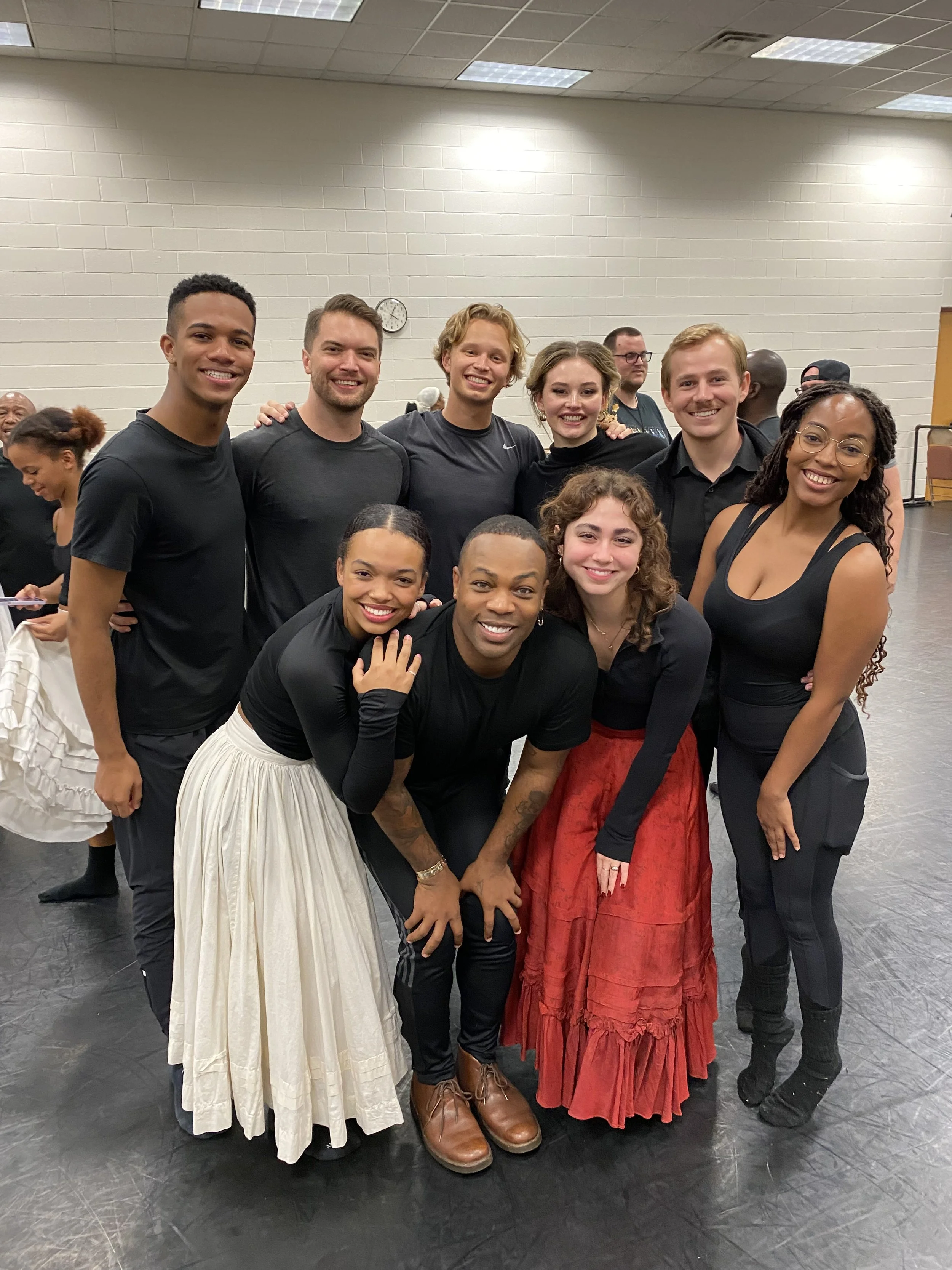 A group of nine diverse young adults posing together in a room with a gray floor and white brick wall, smiling and showing camaraderie.