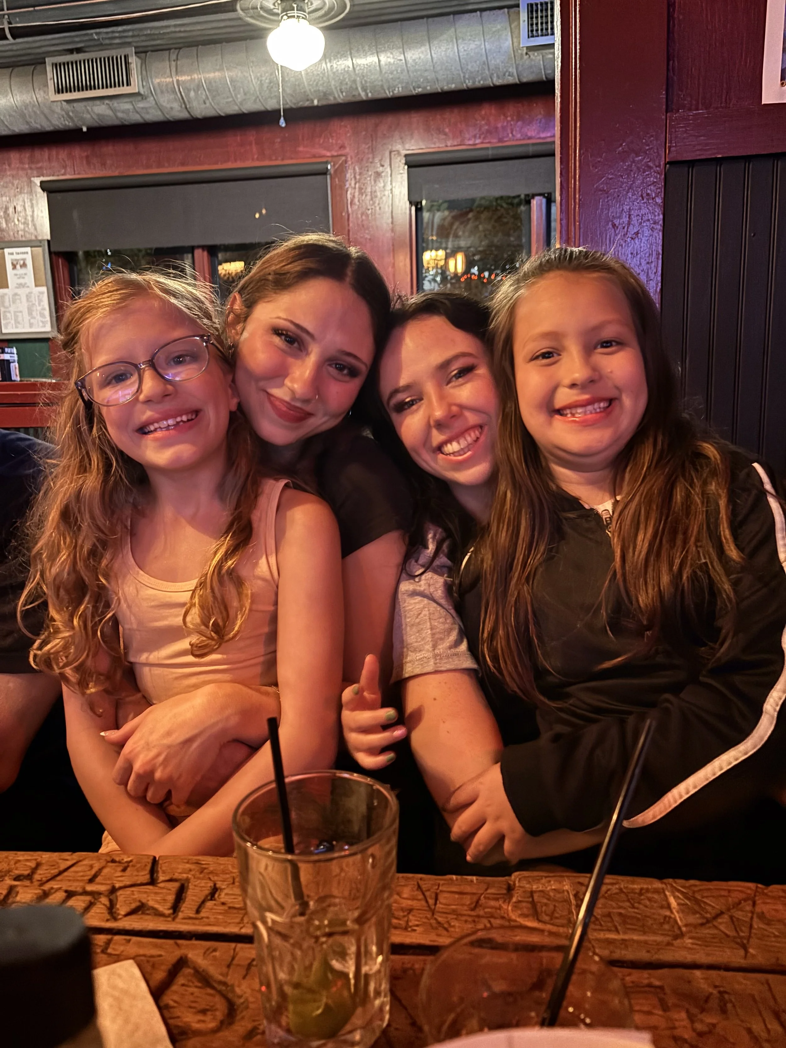 Four women sitting closely together at a restaurant, smiling and posing for a photo indoors with red walls and windows in the background. A drink with a straw is on the table in front of them.