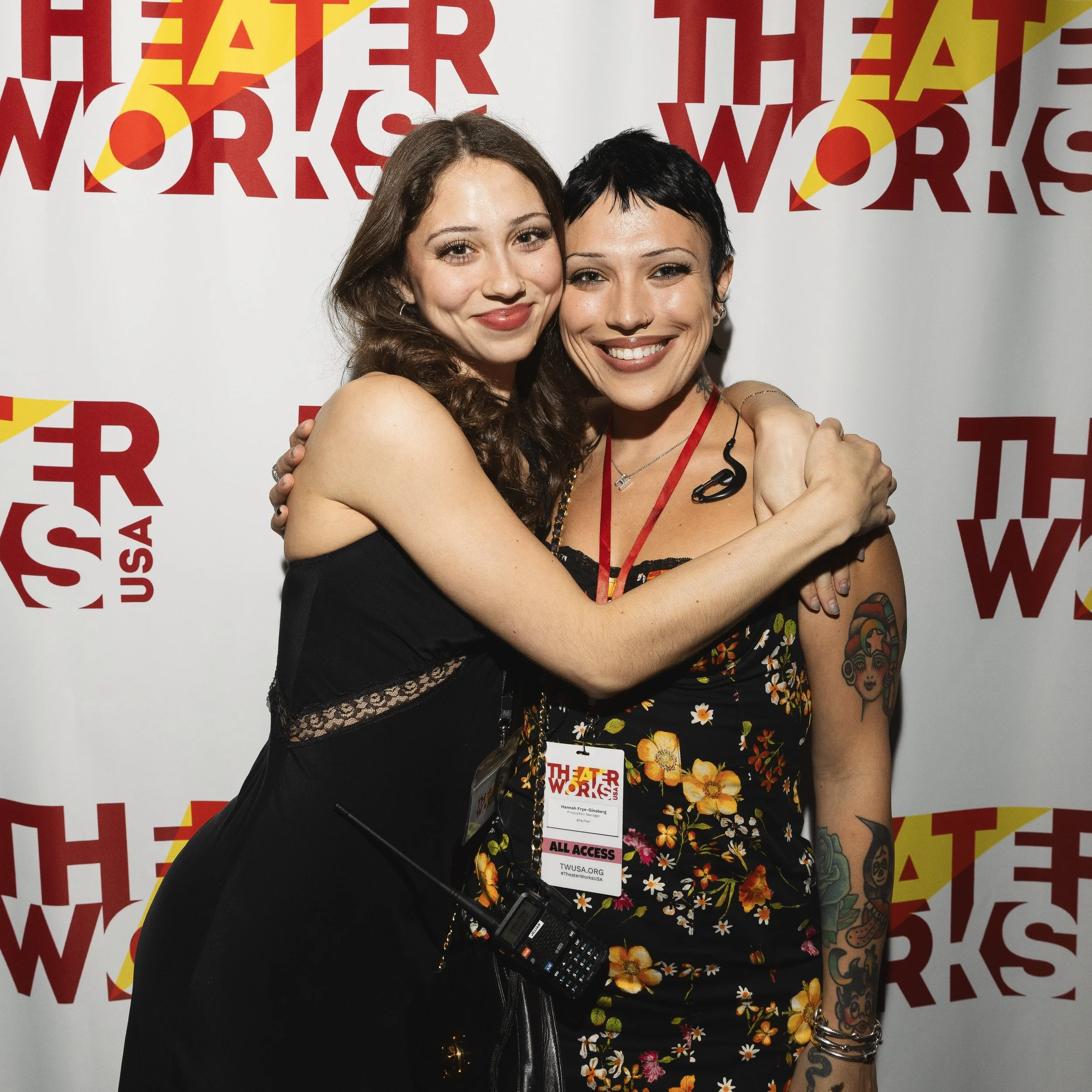 Two women embracing each other and smiling in front of a red and white background with the text 'THEATRE WORKS' repeatedly printed. They are wearing festival passes around their necks, one with a floral dress and the other in black with lace details. The woman in the floral dress has tattoos on her arm.