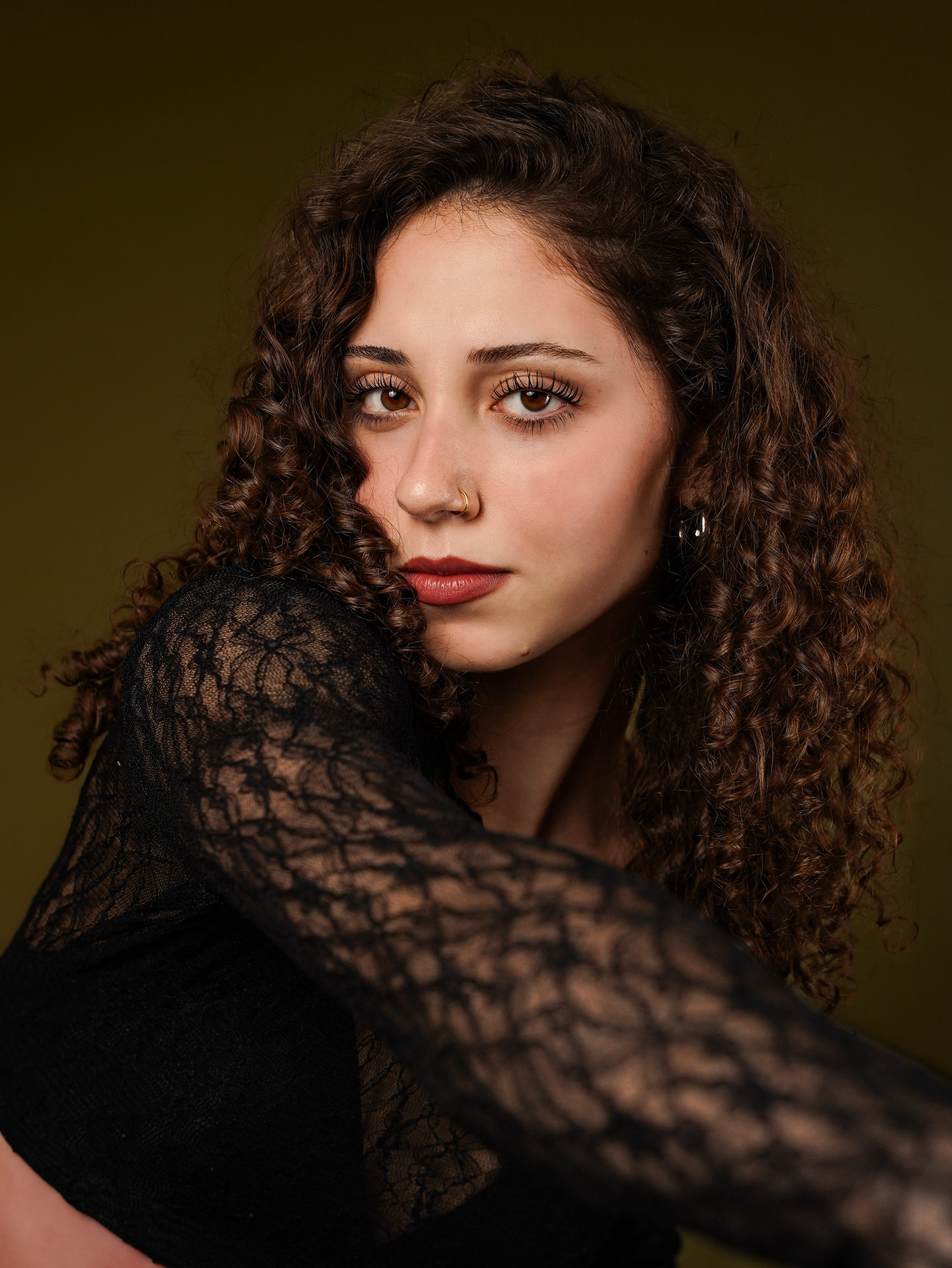 Close-up portrait of a young woman with curly brown hair and brown eyes, wearing a nose ring and a black lace top, against a blurred brown background.