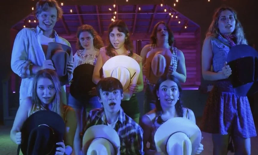 A group of seven young people performing in a theater or on stage with festive string lights overhead. They are holding hats and a large yellow paper with a smiling face, displaying surprised or excited expressions.