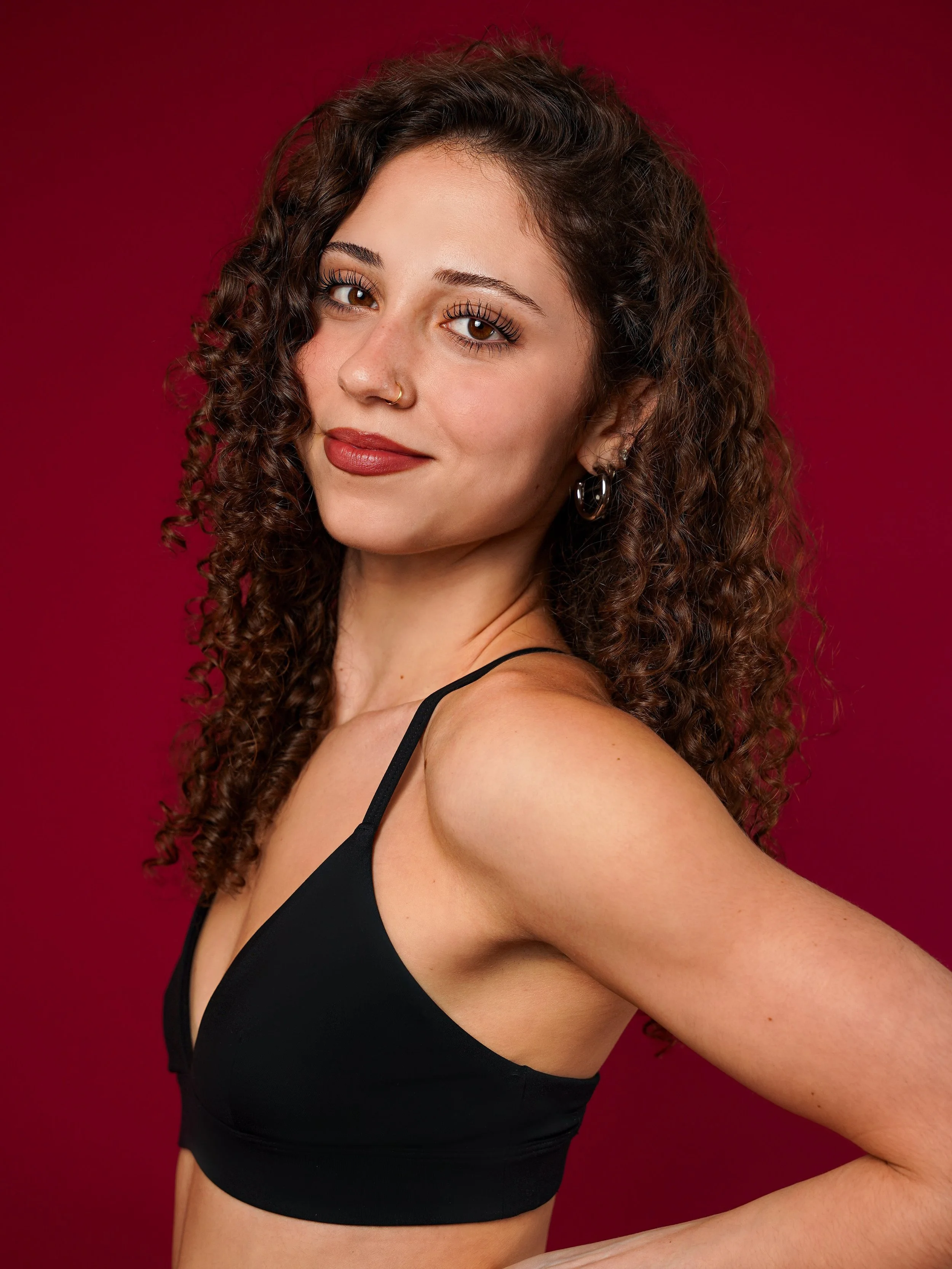 Close-up of a woman with long curly brown hair, brown eyes, wearing makeup, a nose ring, black strap top, against a solid red background.
