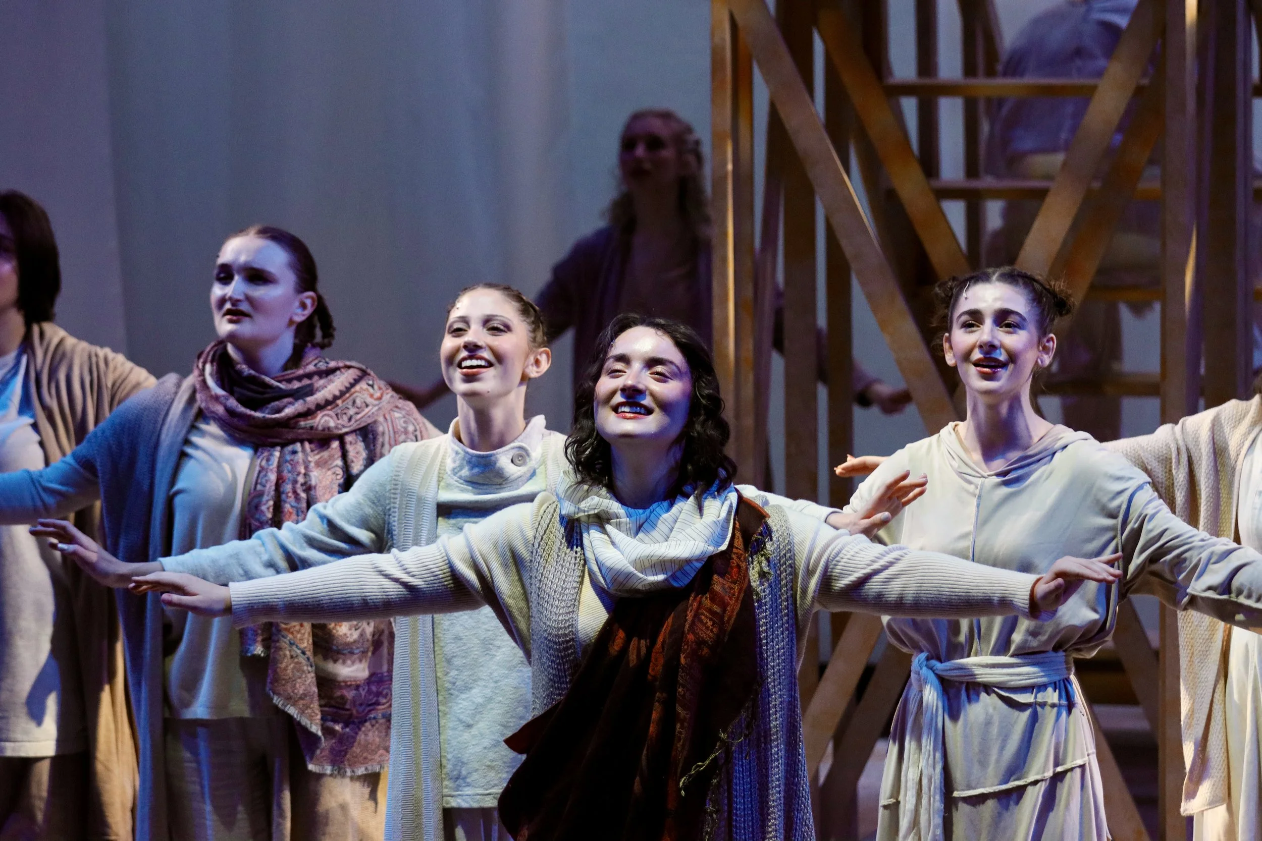 A group of young women dressed in costumes, posing with arms outstretched, on stage during a theatrical performance.