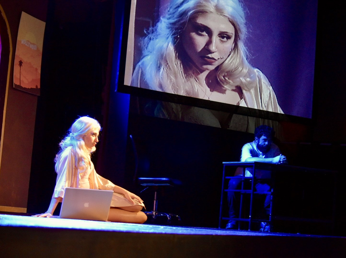 A stage scene with a woman sitting cross-legged on the floor with a laptop in front of her, and a man sitting at a desk. A large screen above displays a close-up of the woman’s face.