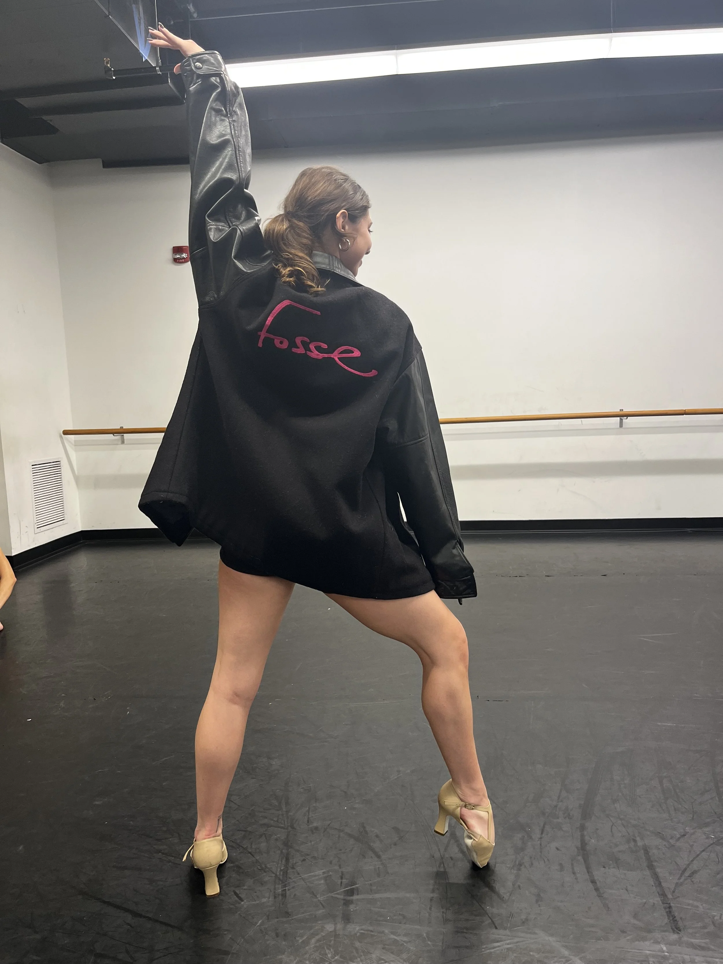 A woman wearing a black jacket with pink embroidery, beige high heels, and performing a dance move with one arm raised and one leg extended, in a dance studio with a white wall and a ballet barre.