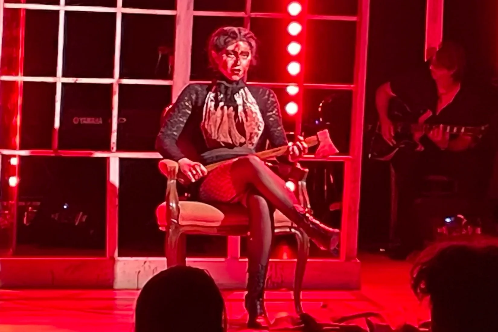 Woman performing on stage with a red background, sitting on a chair, wearing a black lace top, patterned shorts, and high boots, holding a broom, with a musician playing guitar in the background.