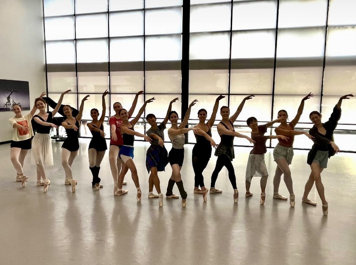 Group of ballet dancers practicing together in a dance studio with large windows in the background.