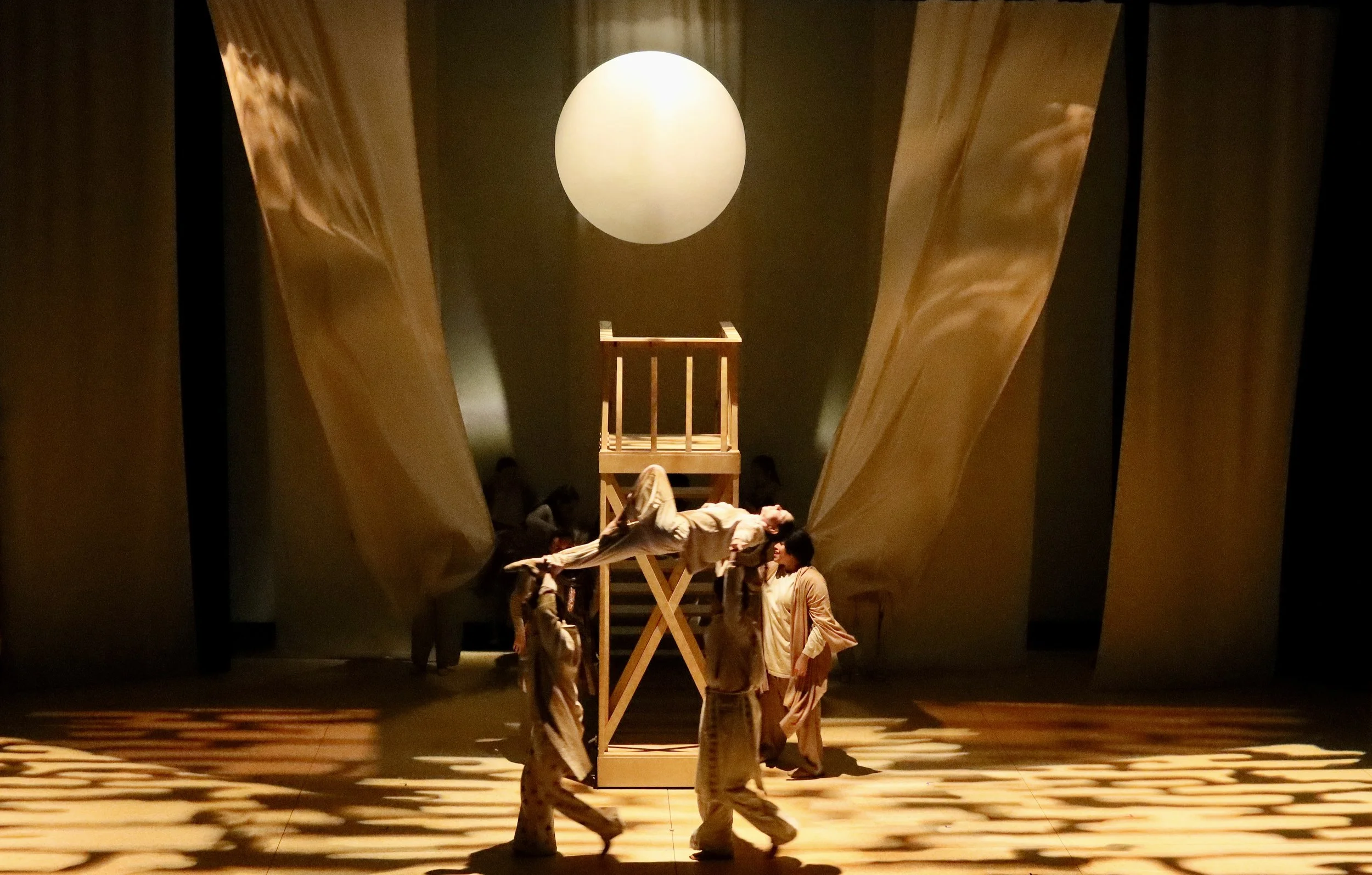 Performers on stage under a large spherical light, with wooden structures and curtains in the background.