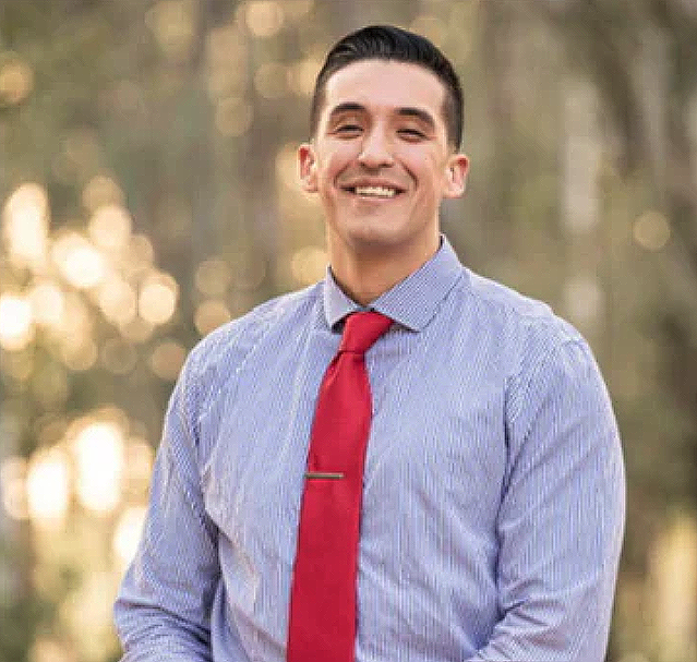 A smiling man in a light blue dress shirt and red tie standing outdoors with blurred trees in the background.