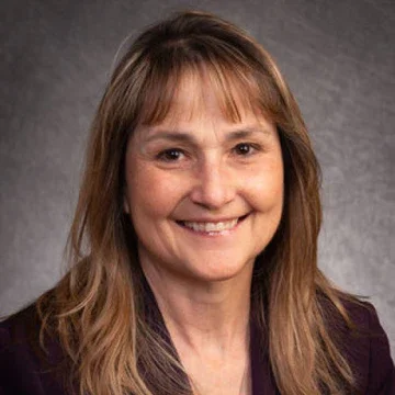 A woman with shoulder-length light brown hair smiling at the camera against a gray background.