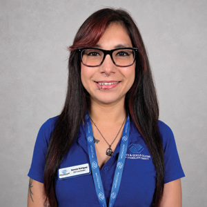 A woman with long dark hair, glasses, and a blue uniform smiling at the camera against a gray background.