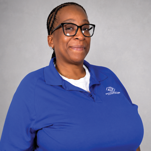 A woman with glasses and braided hair, wearing a blue collared shirt with a logo, standing against a plain grey background.