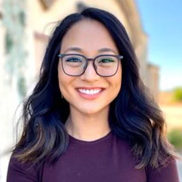 Smiling woman with glasses and dark hair outdoors on a sunny day.