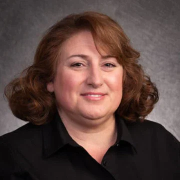 A woman with shoulder-length red hair wearing a black shirt, smiling in front of a gray background.