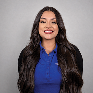 Portrait of a woman with long dark hair, smiling, wearing a blue collared shirt and black cardigan against a light gray background.