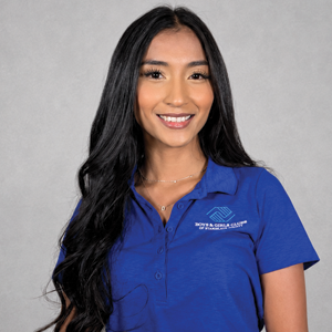 Portrait of a young woman with long black hair, wearing a blue polo shirt with a logo, standing against a plain gray background.