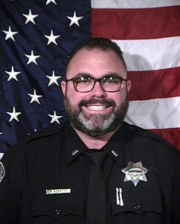 Male police officer with glasses and a beard, smiling, in front of an American flag background.