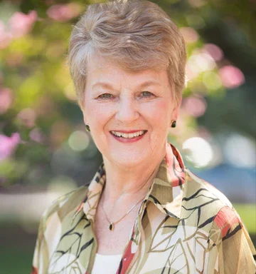 Portrait of an older woman smiling outdoors with pink flowers in the background.