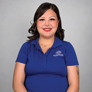A woman with shoulder-length dark hair, wearing a blue polo shirt with a logo, smiling in front of a gray background.